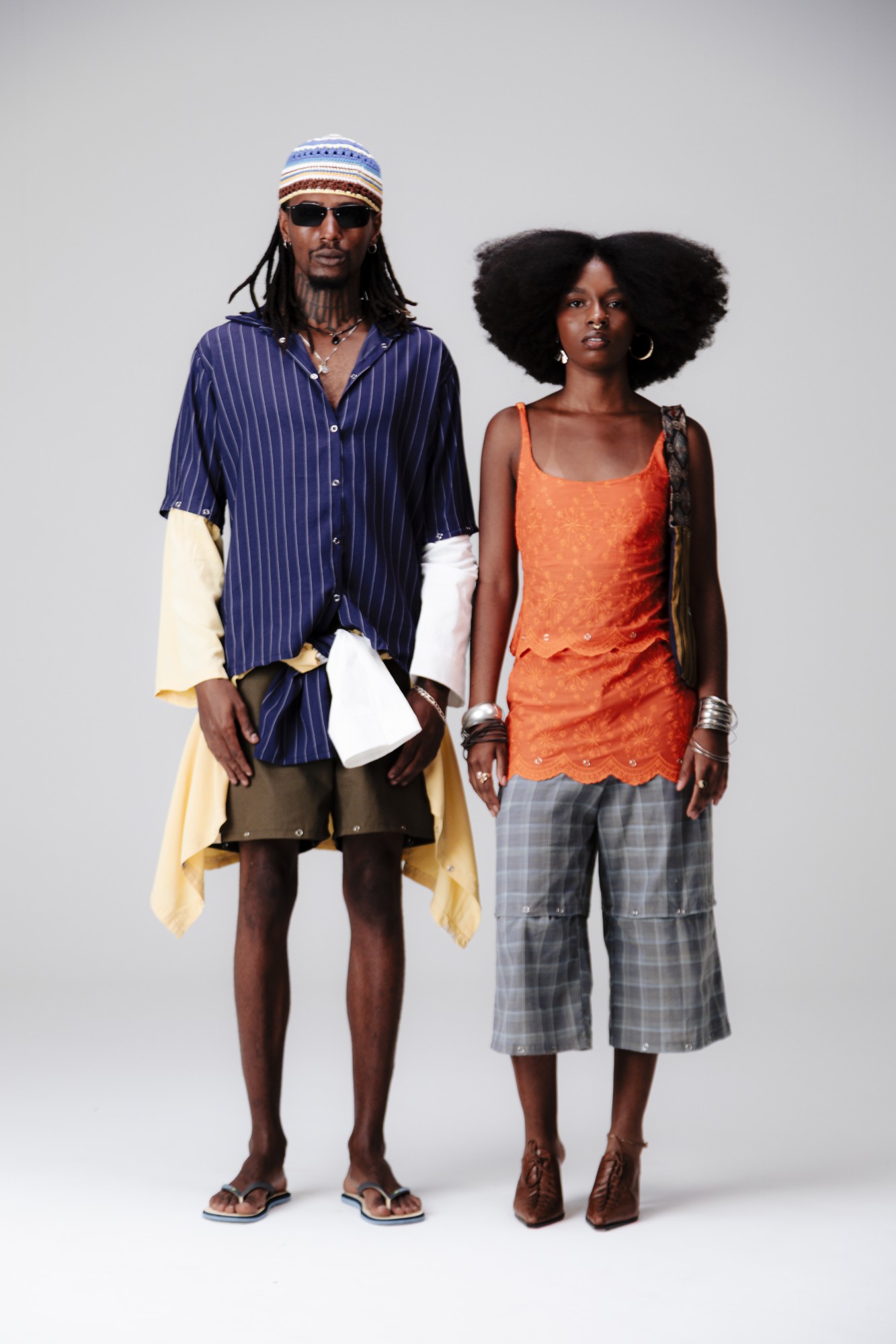 Dupla de modelos em pé: camisa listrada azul-marinho com touca de crochê, e regata laranja bordada com bermuda xadrez cinza