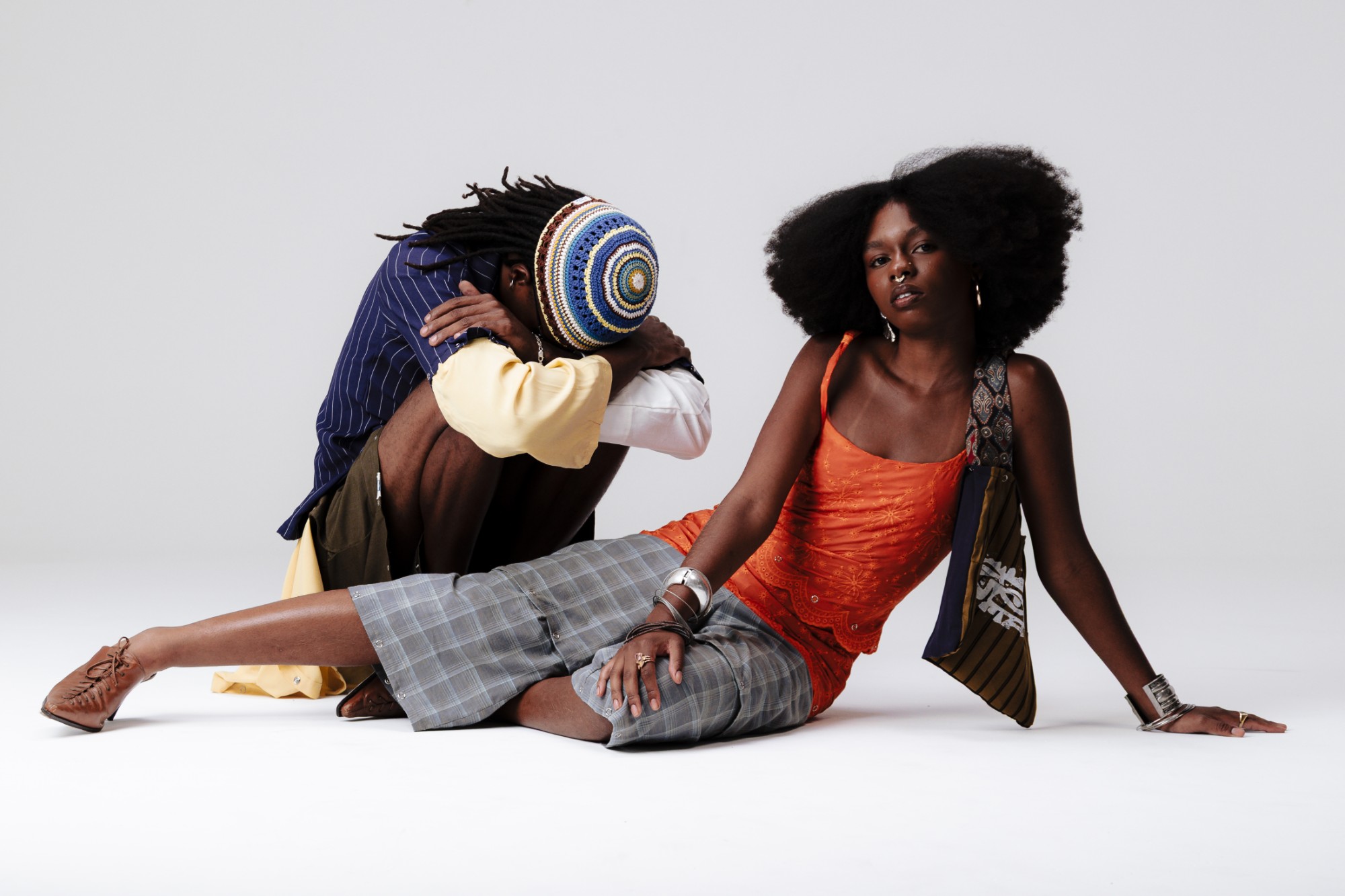 Dupla de modelos no chão: uma pessoa de regata laranja e bermuda xadrez, outra agachada com a touca de crochê
